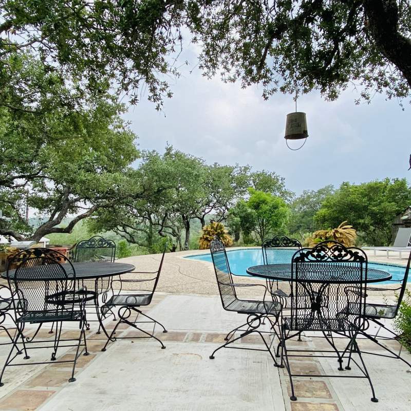 Outside in front of the swimming pool are two, black wrought iron tables each with four chairs for a seating of eight. A repurposed metal bucket light fixture hangs from a tree.